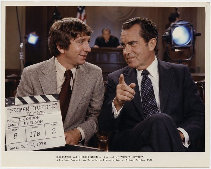 Color production still of Bob Denver and Richard Nixon seated together on a courtroom set during filming of the 1979 television film Frozen Justice, with a blue spotlight visible behind them.