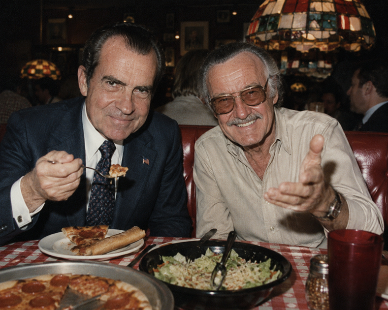 Richard Nixon and Stan Lee sit together at a chain pizza restaurant, smiling at the camera while sharing pizza and salad beneath stained-glass hanging lamps.