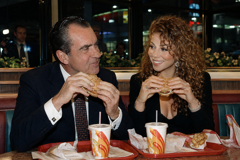 Richard Nixon and Mariah Carey share sandwiches and shakes inside a fast-food restaurant while photographers watch from outside.