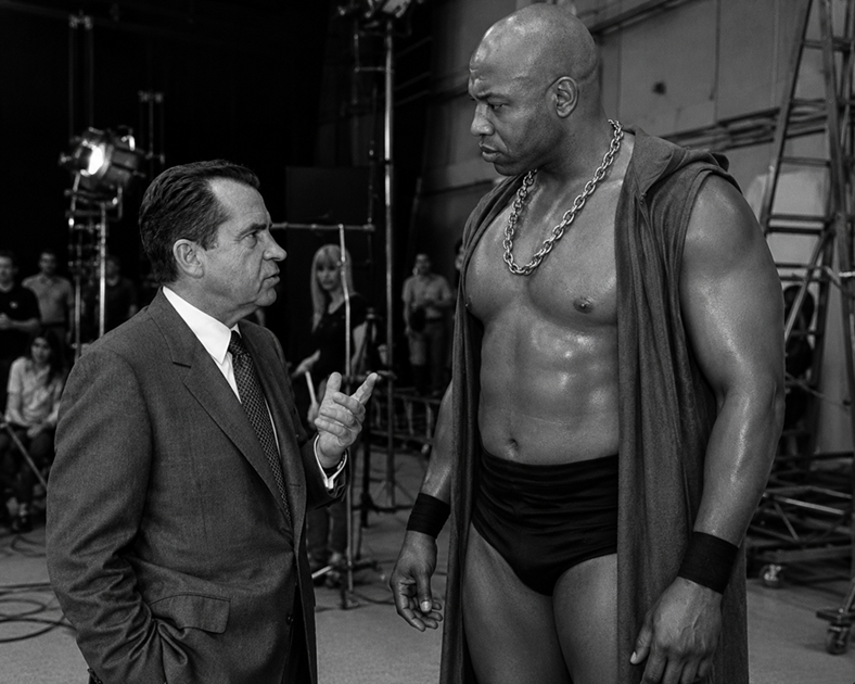 Black-and-white on-set production still depicting Richard “Tricky D” Nixon in a dark suit speaking with towering wrestler and actor Zeus (Tiny Lister) during the filming of No Holds Barred. Nixon gestures calmly while Zeus stands imposingly in costume, towering over him amid studio lighting and film equipment.