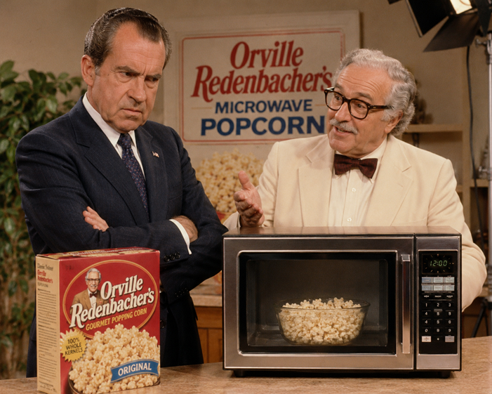 Richard Nixon stands beside Orville Redenbacher on the set of a vintage microwave popcorn commercial. While Redenbacher enthusiastically gestures toward a microwave oven, Nixon folds his arms and eyes the appliance with deep suspicion, as if questioning its strategic implications. A box of Orville Redenbacher popcorn sits on the counter in front of them.